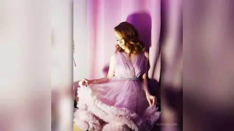 Elegant woman in a flowing lavender tulle gown posing in soft pink studio lighting.