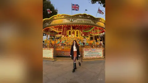 Woman in a black coat and brown skirt standing before a glowing vintage carousel at a festive night carnival.