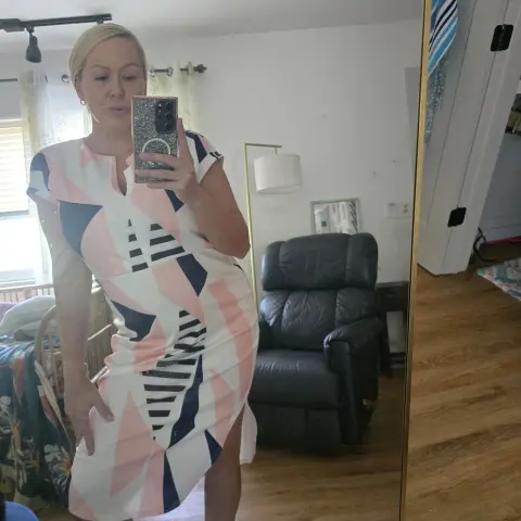 Blonde woman in a geometric print midi dress taking a mirror selfie in a cozy living room.