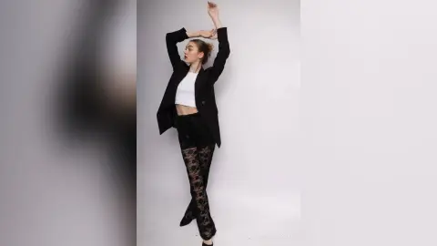 Fashionable woman in a black blazer and lace leggings posing confidently against a white studio background.