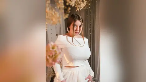Young woman in a white off-the-shoulder outfit posing in a dreamy, floral decorated setting.