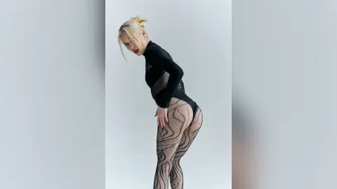 Blonde woman in a black long-sleeve bodysuit and patterned tights posing against a white studio background.