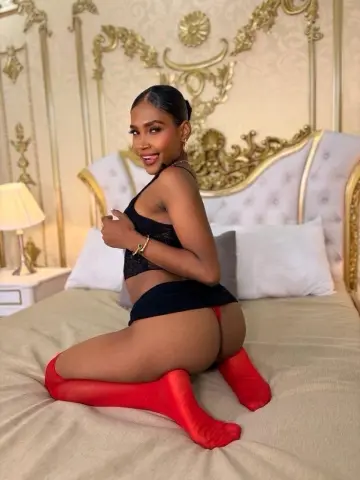 Smiling woman in black lingerie and red socks sitting on a bed in a luxurious gold bedroom.
