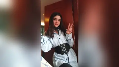 Young woman in a light blue polka dot blouse and black corset belt posing playfully in a room.