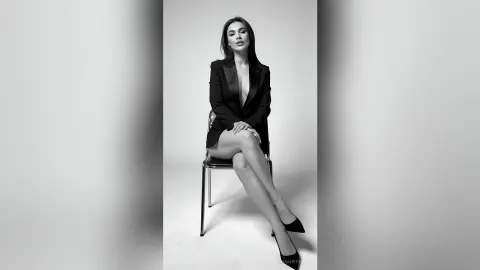Elegant woman in a black blazer and heels posing confidently in a minimalist studio setting.