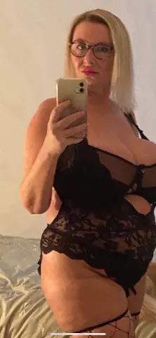 Blonde woman in black lace lingerie bodysuit taking a mirror selfie in a bright bedroom setting.