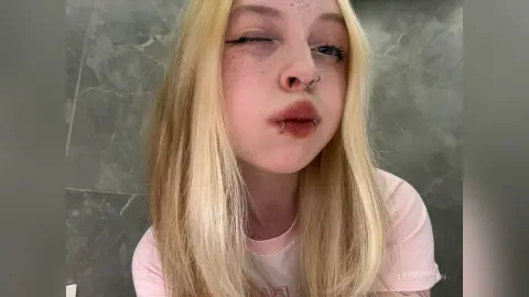 Blonde woman with freckles and lip piercings making a playful face in a modern grey bathroom setting.
