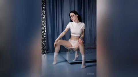 SofiMils poses sitting on a clear bench wearing a white crop top and white undies against a blue curtain background.