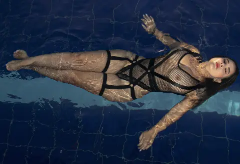 PaolaReyy — PaolaReyy floats on her back in a blue swimming pool wearing a black mesh bodysuit.