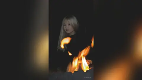 Blonde woman in a black outfit posing behind glowing fire flames in a dark, moody setting.
