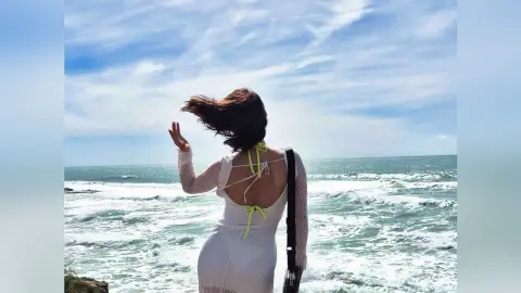 Woman in a white backless summer dress looking at ocean waves under a bright blue sky.