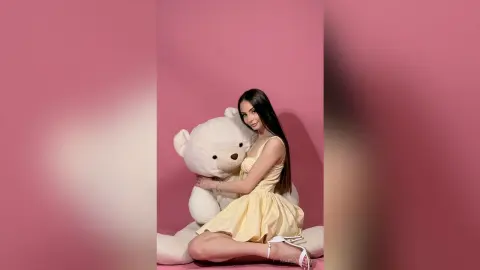 Smiling young woman in a yellow sundress hugging a large white teddy bear against a pink background.