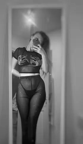 Woman in black sheer crop top and leggings taking a moody black and white mirror selfie.