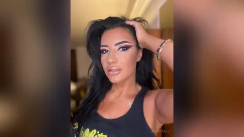 MissyJolie poses with her hand in her long black hair, wearing a black tank top and a silver cross necklace.