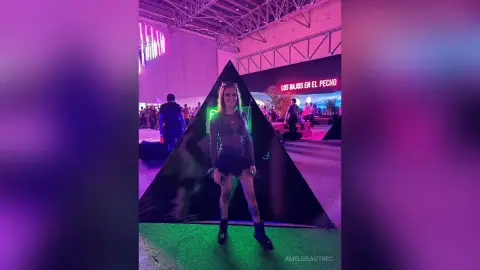 AmelieLautrec stands in front of a black triangular backdrop, wearing a grey long-sleeved top and a short black skirt.