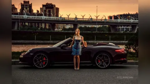 StelaBrown stands next to a dark maroon convertible car wearing a blue denim dress and light-colored heels against a sunset city skyline.