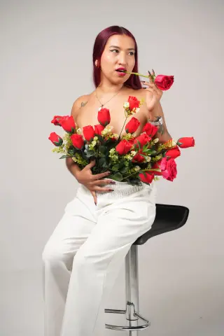 Young woman with red hair in white trousers holding a large bouquet of red roses in a studio setting.