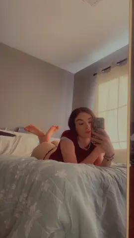 Young woman in a burgundy shirt relaxing on a floral bed in a cozy, sunlit bedroom.