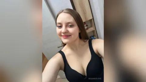 MiaAngelove — MiaAngelove smiles at the camera while wearing a black tank top in a room with wooden shelving.