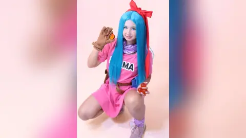 Cheerful girl in pink anime cosplay with long blue hair and red bow posing against a soft pink background.
