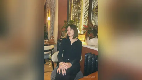 Young woman in a black long-sleeve shirt sitting in a cozy, warm-lit living room with indoor plants.