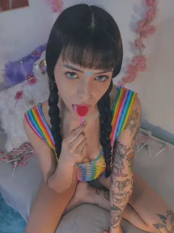 Young woman with dark braids in a rainbow crochet top eating a red lollipop in a dreamy pastel bedroom.