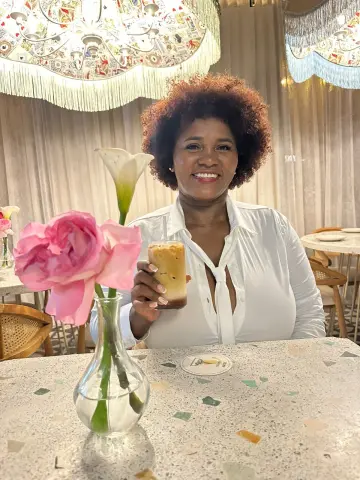 Smiling woman in a white blouse holding a cocktail at a bright cafe table with pink roses.