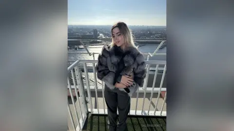 ValerieGracy stands on a balcony wearing a thick grey fur coat and dark trousers against a bright city skyline.