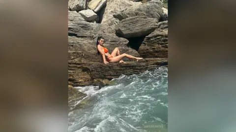 Young woman in an orange bikini relaxing on rocky cliffs by the turquoise ocean waves under bright sunlight.