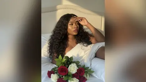 Beautiful woman with long wavy hair relaxing in bed with a red and white rose bouquet in soft sunlight.
