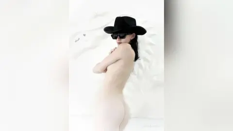 Nude woman in a black wide-brimmed hat and sunglasses posing against a white textured wall in a minimalist style.
