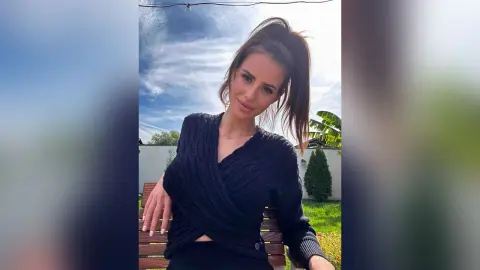 Smiling woman in a black long-sleeve wrap top posing outdoors in a sunny garden setting.