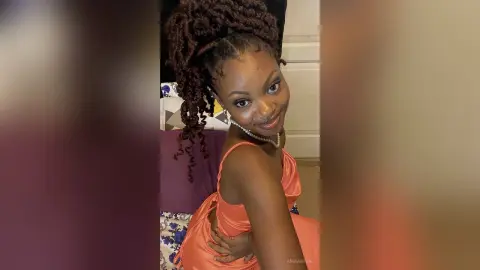 Smiling woman in a peach satin dress with long dark locs posing indoors for a joyful portrait.
