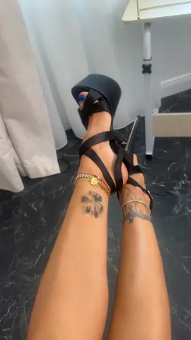 Woman wearing black strappy high heel sandals and gold bracelets with leg tattoos in a modern room.