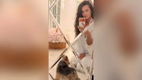 Beautiful woman in white loungewear taking a mirror selfie with her small dog in a bright, floral room.