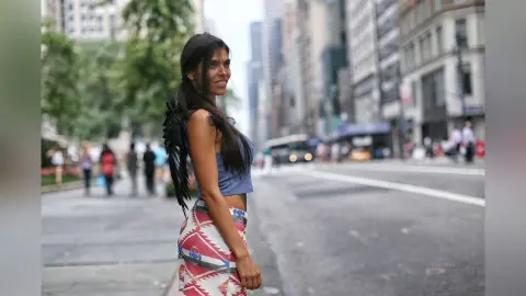 Smiling woman in a blue crop top and patterned skirt wearing black angel wings on a busy city street.
