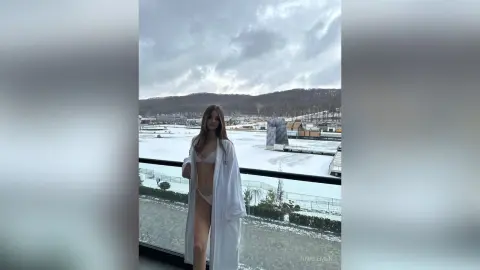 Young woman in a white bathrobe standing on a balcony overlooking a snowy winter landscape.