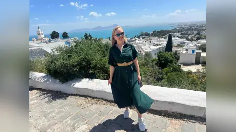 Smiling woman in a dark green midi dress and white sneakers overlooking a sunny Mediterranean coastal town.