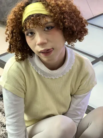 Jessiee_Belle with curly red hair and a yellow headband sticks her tongue out while wearing a pale yellow top.