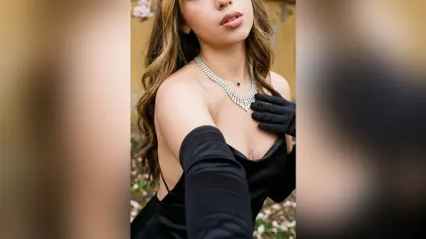 Elegant woman in a black dress and gloves wearing a sparkling diamond necklace in a soft outdoor setting.