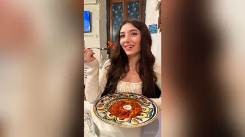 Smiling young woman in a white ribbed top enjoying a delicious pasta dish in a cozy restaurant setting.