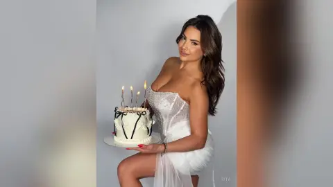 Smiling woman in a white strapless dress holding a lit birthday cake in a bright studio setting.
