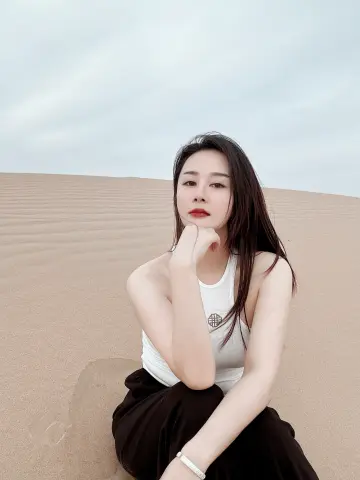 Young woman in a white tank top sitting on sand dunes under a cloudy sky with a serene expression.