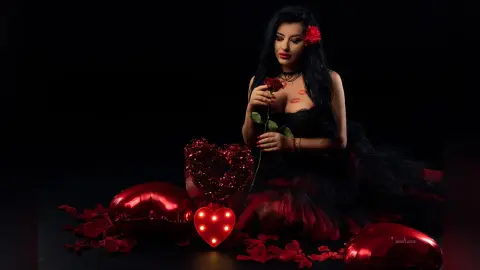 MissyJolie holds a red rose against her chest while wearing a black lace bustier and sitting among red rose petals and heart-shaped decorations.