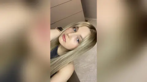 Blonde woman with blue eyes wearing a dark top in a bright, minimalist indoor setting.