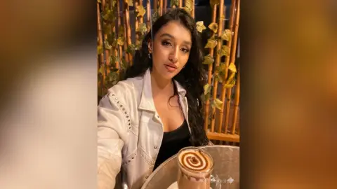 Young woman in a white denim jacket smiling at a cafe with a latte in a cozy evening setting.
