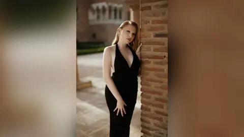 RubySoul poses in a long black dress with a deep v-neck, leaning against a brick wall with her long hair down.
