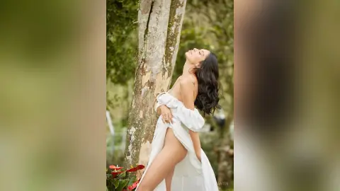 LucyMartin poses outdoors against a tree trunk, wearing a white off-the-shoulder dress that pulls up to reveal her bare thigh and pussy.