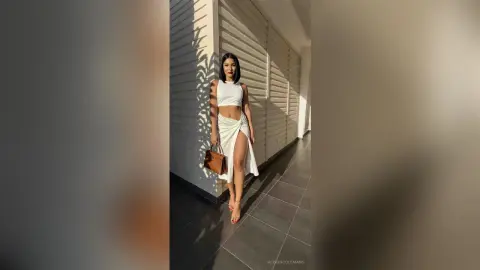 AlisonColemans poses outdoors in a white crop top and long slit skirt, standing against a white slatted wall in the sunlight.
