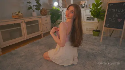 Smiling young woman in a pink camisole and white skirt sitting on a fluffy rug in a cozy, bright bedroom.